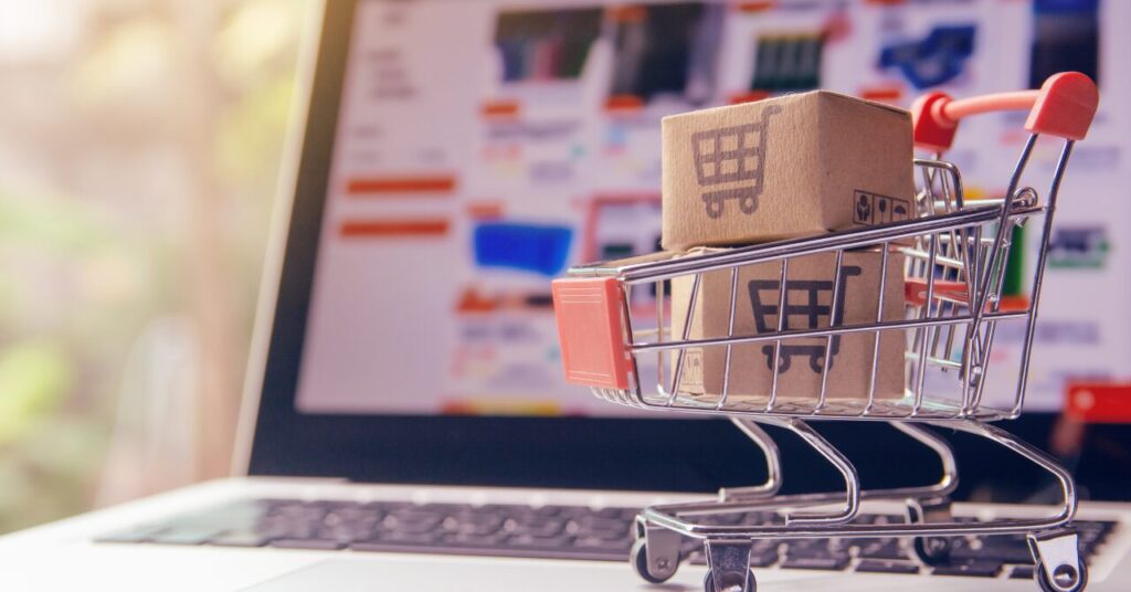 ノートパソコンの前に置かれたショッピングカートと段ボール箱の写真。オンライン購入や価格比較、コスパ検討をイメージした構図。