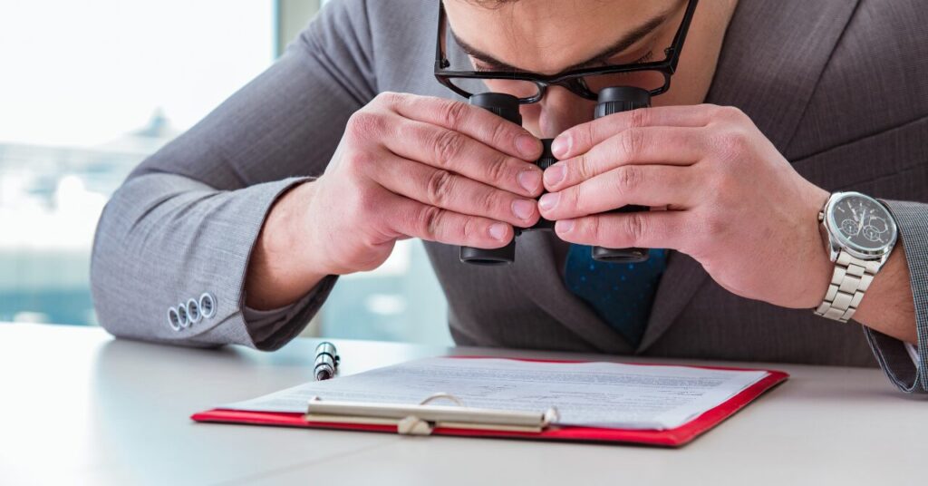 双眼鏡で書類をのぞき込み、細かいポイントを真剣にチェックしている男性のユーモラスな写真。